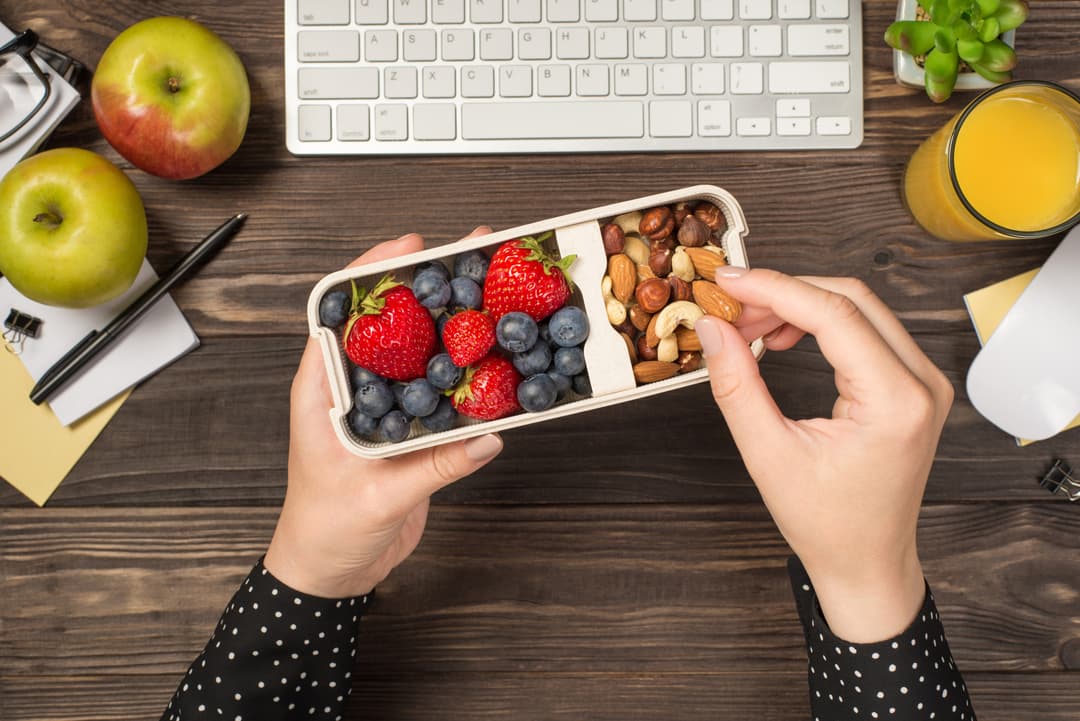 First person top view photo of woman's hands holding lunchbox with healthy meal nuts and berries over apples glass of juice flowerpot stationery keyboard mouse on isolated dark wooden table