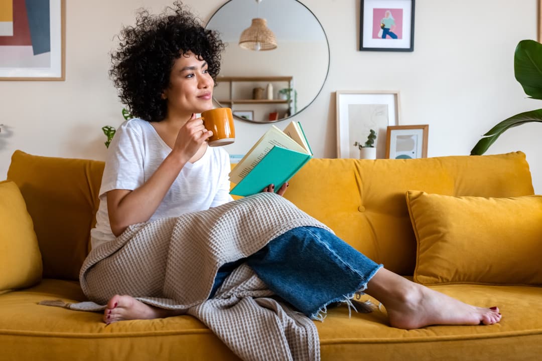 woman reading a book at home, drinking coffee sitting on the couch.