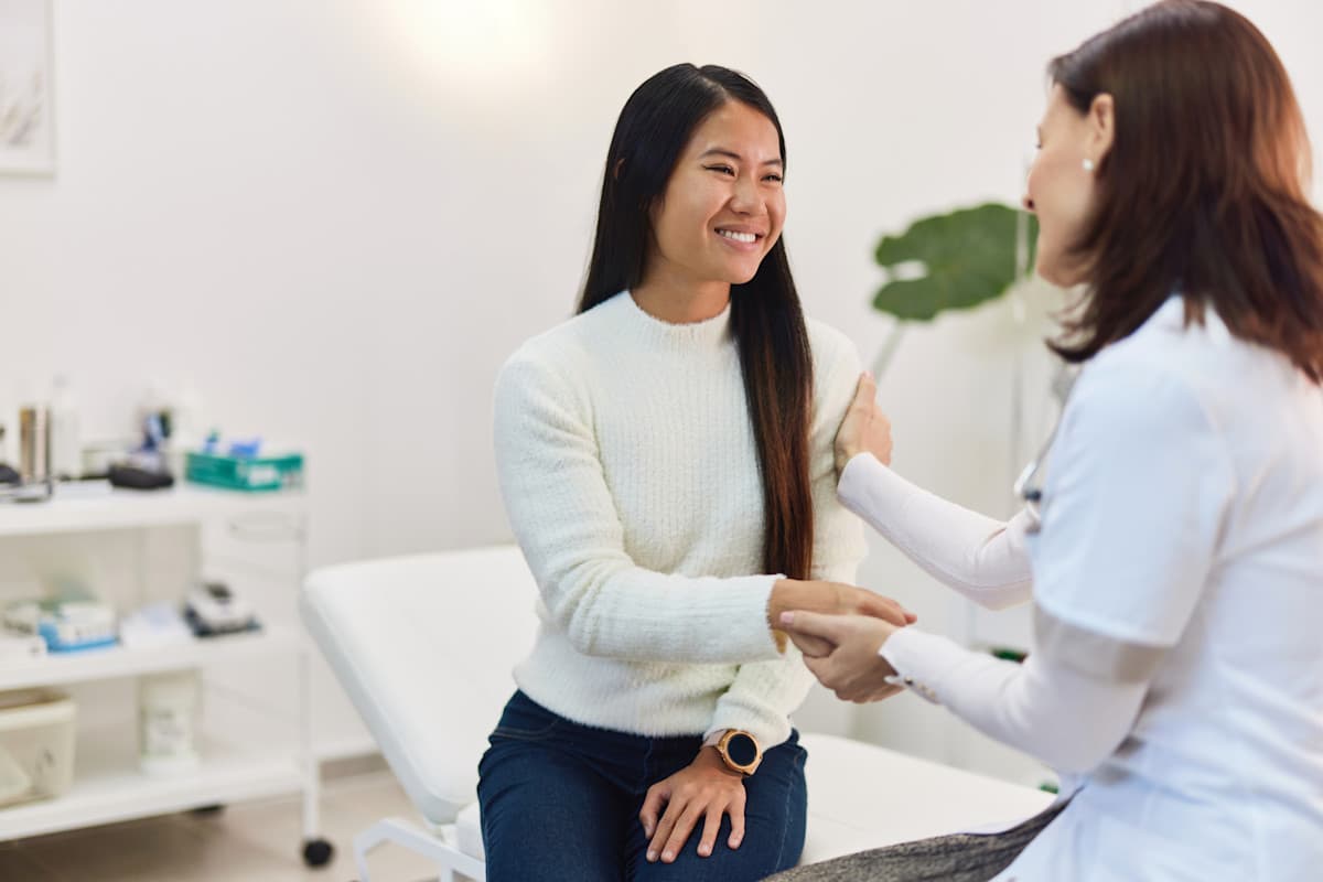 A smiling Asian woman patient sitting with a female doctor in the examination room
