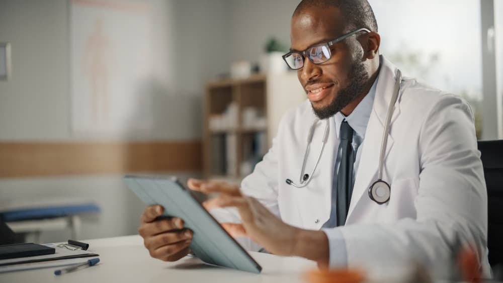 Happy and Smiling African American Male Doctor Wearing White Coat Working on Tablet Computer at His Office.